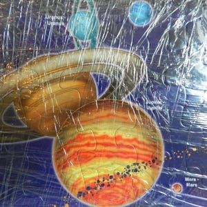 May include: A colorful jigsaw puzzle depicting the solar system. The puzzle features planets like Saturn with its rings, Jupiter, Uranus, Mars, and Pluto. The planets are set against a dark blue background with the planets' names.