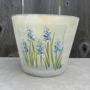 May include: A cream-colored ceramic planter with a hand-painted design of blue flowers and green leaves. The planter has a slightly flared rim and a smooth, textured surface.