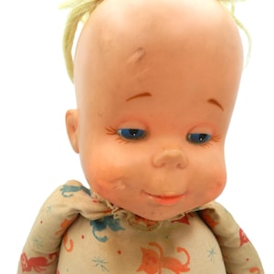 May include: A doll with a smooth, light-toned plastic head, blue eyes, and sparse yellow hair. The doll wears a beige fabric garment featuring a repeating pattern of small orange and blue cats interspersed with tiny red and blue star-like shapes.