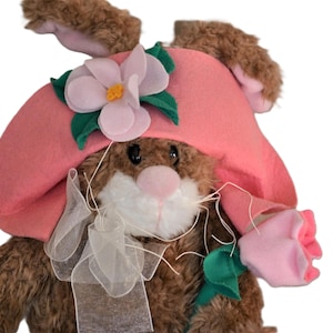 May include: A plush brown bunny wearing a pink hat with a pink flower and green leaves. The bunny is holding a pink flower in its paw.