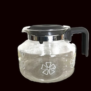May include: A clear glass coffee carafe with a black lid and handle. The carafe has white floral designs and measurement markings. The carafe is designed to hold up to 10 cups of coffee.