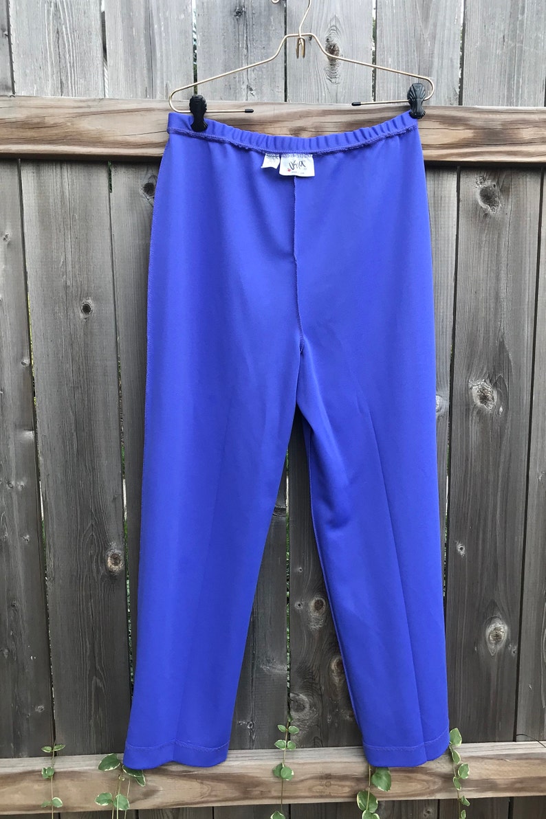 Pode incluir: Uma cal&ccedil;a roxa de perna reta e cintura larga. A cal&ccedil;a est&aacute; pendurada em um cabide dourado contra um fundo de madeira.