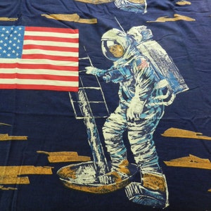 May include: A blue fabric with a repeated pattern of an astronaut standing on the moon, an American flag, and a gold-colored moon surface.
