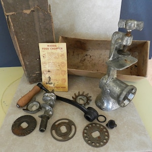 May include: Vintage Wards Food Chopper in its original box with instructions. The metal food chopper has a crank handle and various attachments for grinding meat, vegetables, and nuts. The box is made of cardboard and has a brown and white color scheme.