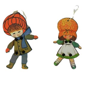 May include: Two vintage wooden ornaments depicting children in winter attire. One figure wears a large orange hat and a blue scarf, the other a similar hat and a white dress with green trim. Both have black button details.