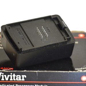 May include: A black Vivitar Dedicated Processor Module, with a rectangular shape and silver pins, sits atop its original box. The box is black with red and white text, including the Canon and Vivitar brand names.