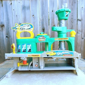 May include: A green and gray plastic toy airport playset with a control tower, a hangar, and a runway. The playset has a yellow sign that says "Sky Station Jet Port" and another sign that says "Sky Station Hangar".