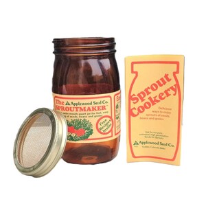 May include: A brown glass jar with a white label that reads "The Applewood Seed Co. SPROUTMAKER" and a metal lid with a mesh screen. The jar is next to a yellow card with the text "Sprout Cookery" in red and black.