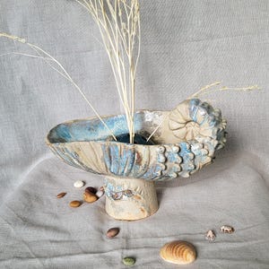 Pode incluir: Uma taça de cerâmica sobre pedestal com um design semelhante a uma concha e um esmalte texturizado em azul e bege. A taça tem um suporte floral escuro no centro que segura hastes brancas secas. Pequenas pedras e uma concha estão próximas.
