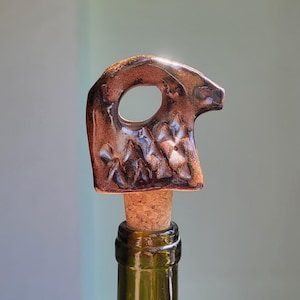 May include: A decorative bottle stopper with a unique design. The stopper features a brown, metallic-glazed ceramic top with an oval opening and textured details. It is attached to a cork, and sits atop a green glass bottle.