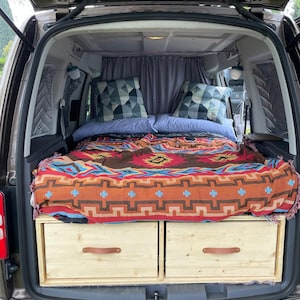 Puede incluir: La parte trasera de una furgoneta con una plataforma de cama y dos cajones de madera con tiradores de cuero. La cama está cubierta con una manta colorida con un patrón geométrico.