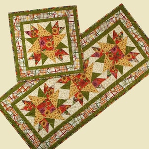 Peut inclure: Un ensemble de trois chemins de table matelassés sur le thème de l'automne. Les chemins de table présentent un motif en étoile éclatante dans des tons de vert, orange et jaune avec un imprimé floral. Les chemins de table sont bordés d'un tissu à carreaux vert et blanc.