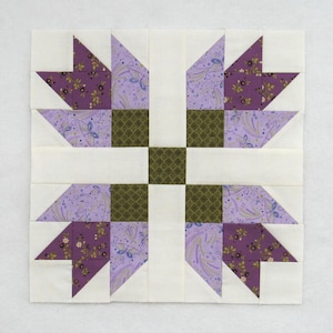 May include: A square quilt block featuring a symmetrical design. The block incorporates white, lavender, and dark green fabrics. The design includes stylized leaf shapes and geometric patterns, creating a visually balanced composition.