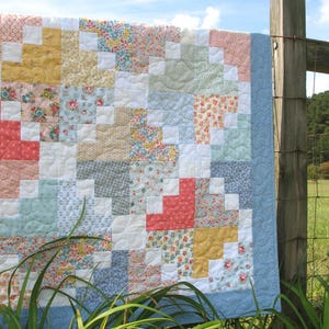 May include: A handmade quilt with a patchwork design, featuring squares of various floral and patterned fabrics in pastel colors. The quilt has a light blue border and is hanging on a wooden fence, with green grass and a blue sky in the background.