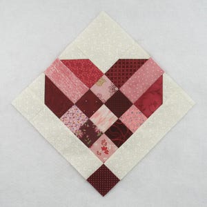 Puede incluir: Un bloque de colcha de retazos en forma de corazón, hecho con varios tonos de tela roja y rosa. El bloque está rodeado de una tela blanca con un patrón sutil.