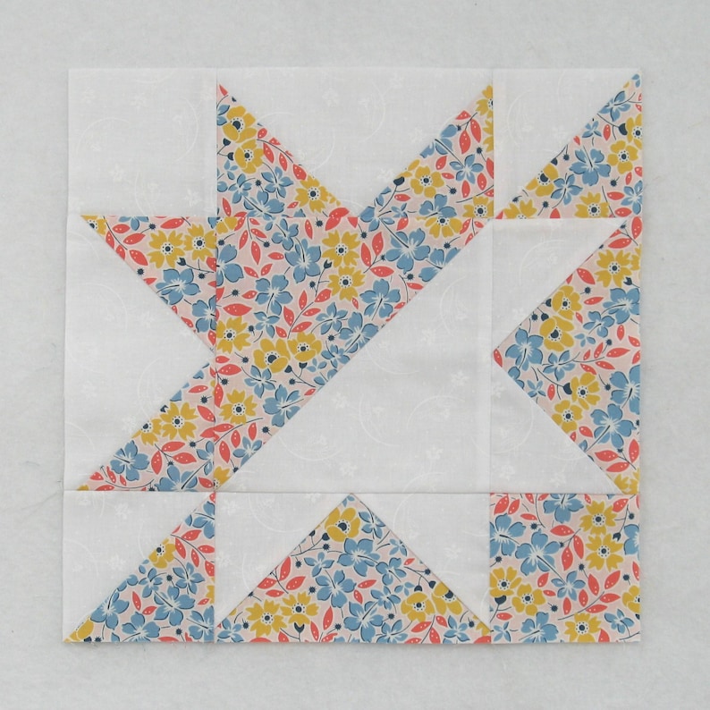 May include: A quilt block featuring a star design, crafted from fabric scraps. The star is made from white fabric with a floral print in shades of blue, yellow, and pink. The background is white.