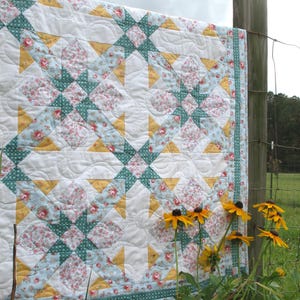 May include: A white quilt with a patchwork design featuring yellow, green, and blue floral fabric. The quilt is hanging on a wooden fence post with a field in the background.