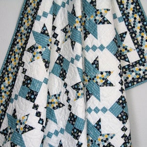May include: A white quilt with a blue, black and yellow star pattern. The quilt is made with a variety of fabrics, including a floral print and a geometric print. The quilt is folded in half and is hanging on a white wall.