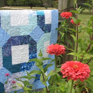 Puede incluir: Una colcha de retazos azul y blanca con un diseño geométrico cuelga de un poste de madera de una valla. La colcha está hecha de varios tonos de tela azul y blanca. La colcha está rodeada de flores rosas.