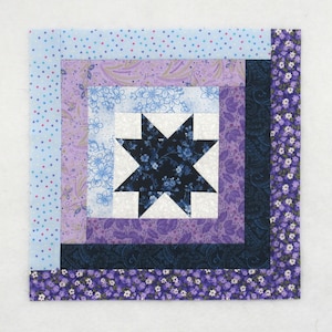 May include: A square quilt block featuring a central star design in navy blue floral fabric, surrounded by concentric rectangles of various patterned fabrics in shades of purple, blue, and white. The block is bordered with a floral print.