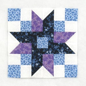 May include: A square quilt block with a star pattern. The star is made from purple, black, and blue floral fabric. White squares and blue floral fabric complete the design. This patchwork design is suitable for quilting projects.