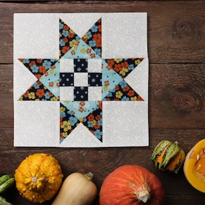 Könnte beinhalten: Ein weißer Stoffquadrat mit einem Patchwork-Muster, das einen großen Stern in der Mitte zeigt. Der Stern besteht aus blauem, schwarzem und orangefarbenem Stoff mit einem Blumenmuster. Der Hintergrundstoff ist weiß mit einem dezenten Muster. Das Quadrat ist von verschiedenen Kürbissen und Kürbissen umgeben.
