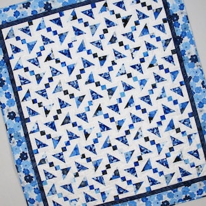 Op de afbeelding: Een blauwe en witte quilt met een geometrisch patroon van driehoeken en vierkanten. De quilt is gemaakt van verschillende blauwe stoffen, waaronder bloemenprints en effen kleuren. De quilt is afgewerkt met een blauwe en witte rand.