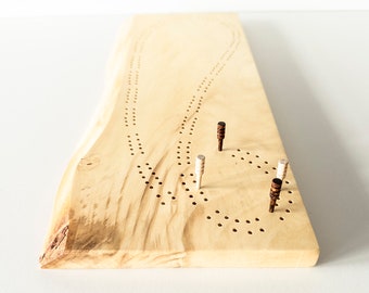 May include: A wooden cribbage board with a natural wood grain finish. The board has multiple rows of small holes and includes four wooden pegs in various colors. The board is rectangular with a live edge on one side.