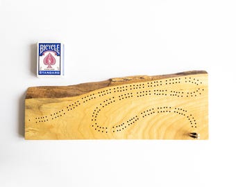 May include: A wooden cribbage board with a natural edge and a deck of Bicycle playing cards. The board is light-colored with a series of drilled holes for scoring. Two metal pegs are inserted at the end of the board. The Bicycle deck is blue and white.