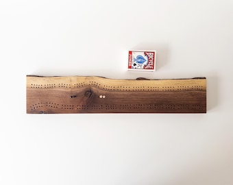 May include: A handcrafted wooden cribbage board with a natural edge, featuring multiple rows of drilled holes for pegging. A standard deck of Bicycle playing cards rests on the board. The wood has a rich, dark brown color.