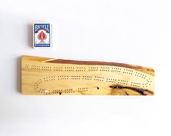 May include: A wooden cribbage board with a natural edge and drilled holes for pegs. A deck of Bicycle playing cards is next to the board. The board is made of light-colored wood with darker brown accents.