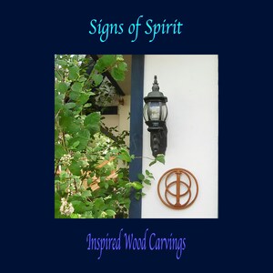 May include: A wooden carving of a symbol with two overlapping circles and a vertical line in the centre. The carving is brown and is mounted on a white wall. The carving is surrounded by green leaves and a black light fixture.