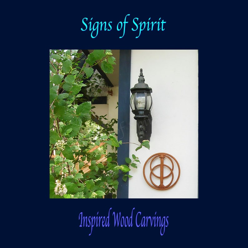 May include: A wooden carving of a symbol with two overlapping circles and a vertical line in the centre. The carving is brown and is mounted on a white wall. The carving is surrounded by green leaves and a black light fixture.