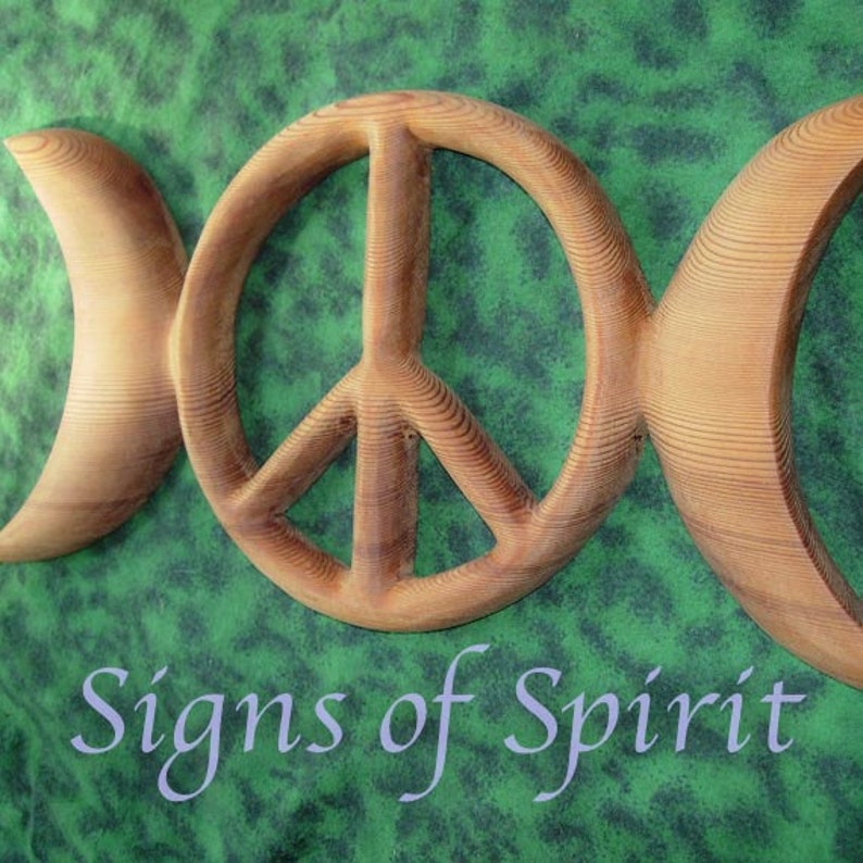 May include: A wooden wall hanging with a triple moon symbol and a peace sign in the center. The wood is a light brown color and the background is a green fabric.