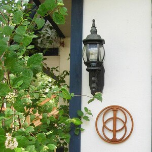 May include: A wooden wall decoration with a brown, circular design featuring a stylised tree with three circles. The decoration is mounted on a white wall next to a black outdoor light fixture.