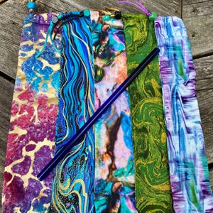 May include: Five colorful fabric pouches with drawstring closures, each containing a blue glass drinking straw. The pouches feature various abstract patterns in shades of purple, blue, green, and gold.