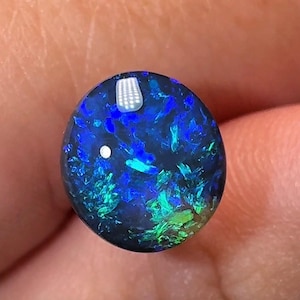 Pode incluir: Uma pedra preciosa de opala negra polida de formato redondo, apresentando uma cor base escura com flashes iridescentes vibrantes de azul elétrico e verde brilhante em toda a superfície, com um corte cabochão liso e arredondado.
