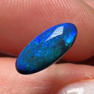 Può includere: Una gemma di opale ovale e lucidata. L'opale mostra un vibrante gioco di colori, con blu e verdi profondi che si muovono all'interno. La superficie riflette la luce, creando un effetto scintillante. La gemma è tenuta tra le dita.