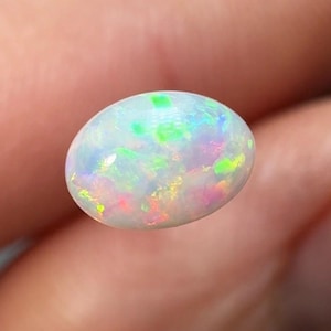 May include: An oval opal gemstone, displaying a white base with flashes of colour including green, blue, pink, and yellow. The polished stone reflects light, showing a spectrum of hues. The gemstone is held between fingers.