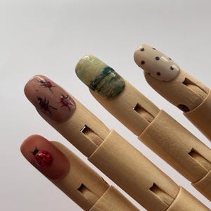 May include: Close-up of four painted fingernails on a wooden hand. The nails feature various designs, including a ladybug, a landscape, a dotted pattern, and an insect. The nails are painted in shades of brown, green, and beige.