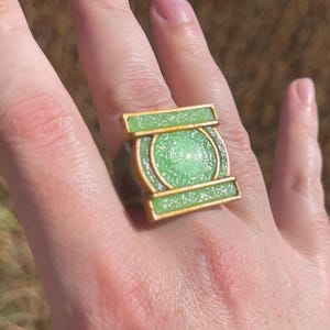 May include: A ring featuring a square-shaped gold-toned frame with a central circular element and two horizontal bars. The design is filled with a bright, glittery lime green material that catches the light, creating a textured, sparkling appearance.