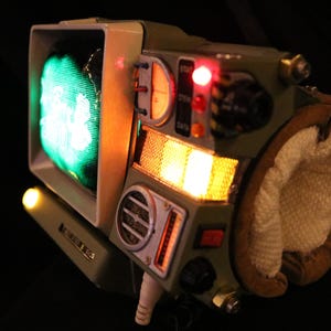 Op de afbeelding: Een uniek, retro-futuristisch apparaat met een klein scherm dat een groen beeld weergeeft. Het apparaat heeft olijfgroene en beige tinten, met diverse knoppen, schakelaars en oplichtende oranje en rode lichten. Een bruin en wit gewatteerd gedeelte is bevestigd.