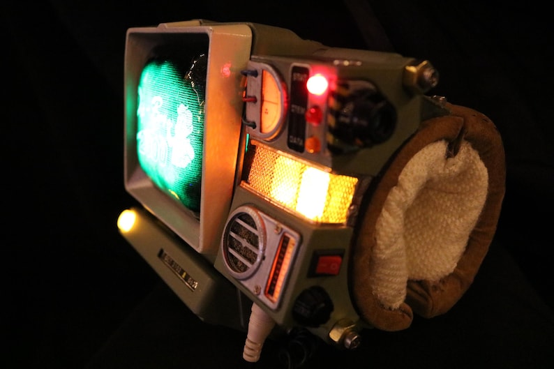 Op de afbeelding: Een uniek, retro-futuristisch apparaat met een klein scherm dat een groen beeld weergeeft. Het apparaat heeft olijfgroene en beige tinten, met diverse knoppen, schakelaars en oplichtende oranje en rode lichten. Een bruin en wit gewatteerd gedeelte is bevestigd.