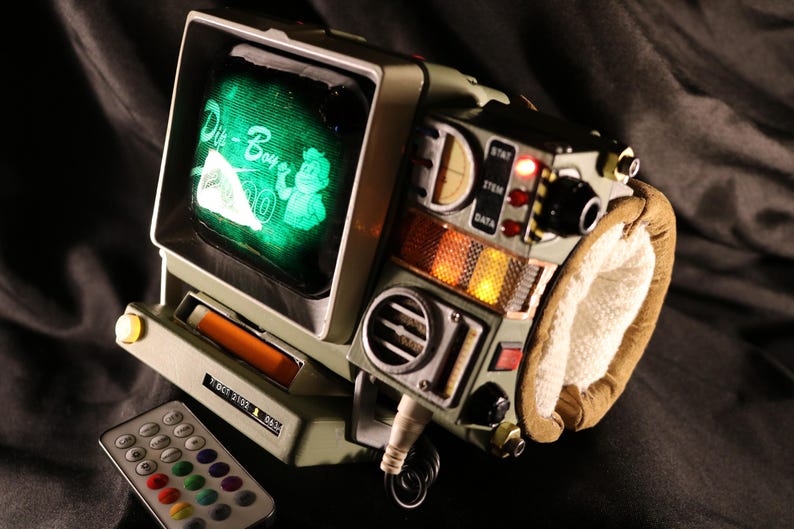 Op de afbeelding: Een retro-stijl apparaat met een groen scherm met "Pip-Boy" graphics. Het apparaat heeft diverse knoppen, lampjes en een polsband. Een afstandsbediening met veelkleurige knoppen ligt in de buurt. Het apparaat is een rekwisiet.