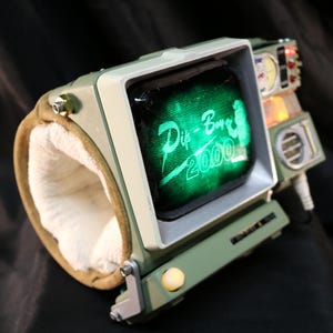 Op de afbeelding: Een retro-futuristisch polsgebonden apparaat met een klein scherm met "Pip-Boy 2000" in het groen. Het apparaat heeft een groen en beige kleurenschema, met een gewatteerde polsband en diverse knoppen. Een uniek rekwisiet.