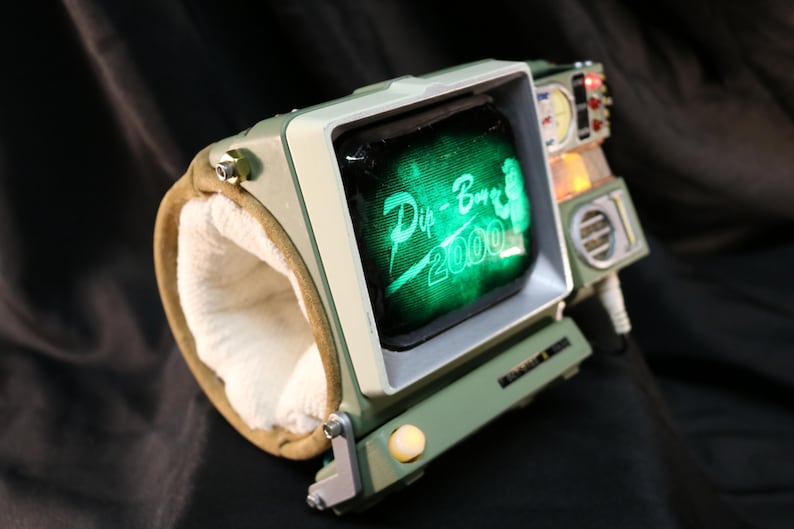 Op de afbeelding: Een retro-futuristisch polsgebonden apparaat met een klein scherm met "Pip-Boy 2000" in het groen. Het apparaat heeft een groen en beige kleurenschema, met een gewatteerde polsband en diverse knoppen. Een uniek rekwisiet.