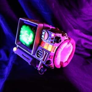 Op de afbeelding: Een retro-futuristisch apparaat met een klein scherm dat een groen beeld weergeeft. Het apparaat heeft een zilveren frame, een roze gevoerde handgreep en diverse knoppen en schakelaars. De sc&egrave;ne is verlicht met paarse en blauwe tinten.