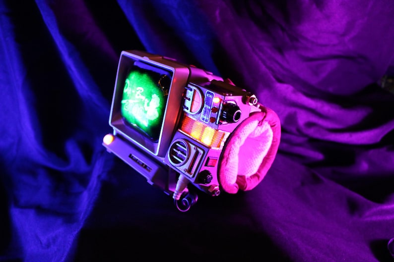 Op de afbeelding: Een retro-futuristisch apparaat met een klein scherm dat een groen beeld weergeeft. Het apparaat heeft een zilveren frame, een roze gevoerde handgreep en diverse knoppen en schakelaars. De sc&egrave;ne is verlicht met paarse en blauwe tinten.