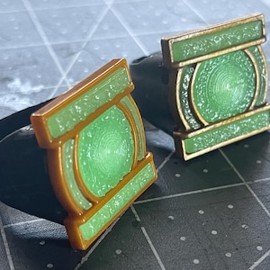 Op de afbeelding: Twee ringen met een vierkant voorvlak en een centraal rond embleem. De ringen hebben een zwarte band, een goudkleurig metalen frame en een getextureerde lichtgroene inleg die de ronde en rechthoekige delen van het ontwerp opvult.