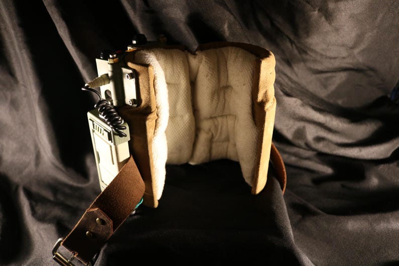 Op de afbeelding: Een bruin en beige rekwisiet met een bruine leren riem en een zwart snoer. Het apparaat heeft een gewatteerd interieur en een metalen component met schroeven. De achtergrond is een donkere, getextureerde stof.